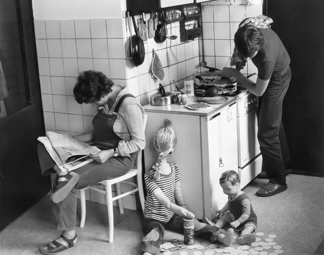Eine Frau liest auf einem Stuhl eine Zeitung, während ein Mann am Herd kocht; zwei als Kinder verkleidete Schaufensterpuppen sitzen auf dem Boden und spielen mit runden Gegenständen, die effiziente Organisation im Haushalt widerspiegeln.