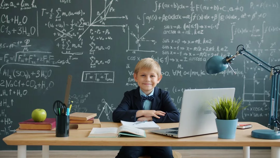Ein junges Kind im Anzug sitzt an einem Schreibtisch mit Büchern und Laptop vor einer Tafel voller Matheformeln und Naturwissenschaften - ein Bild frühzeitiger Förderung.