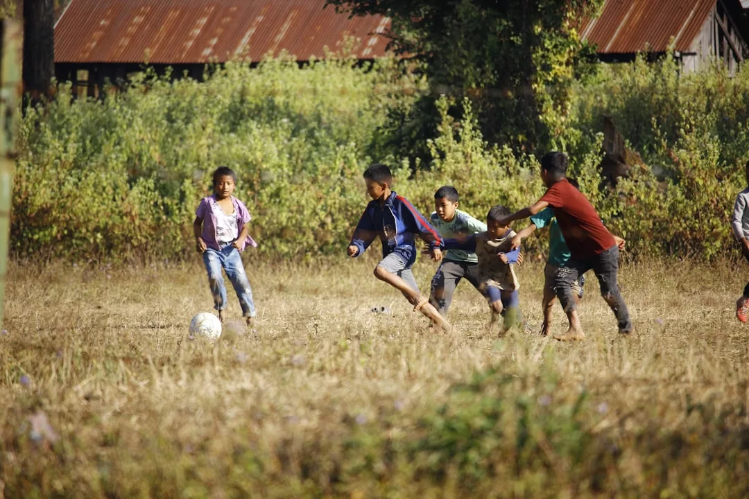 Eine Gruppe von Kindern genießt die körperliche Aktivität, während sie auf einer Wiese Fußball spielen, einige laufen barfuß. Im Hintergrund sind Büsche und ein verrostetes Gebäude mit einem Metalldach zu sehen.