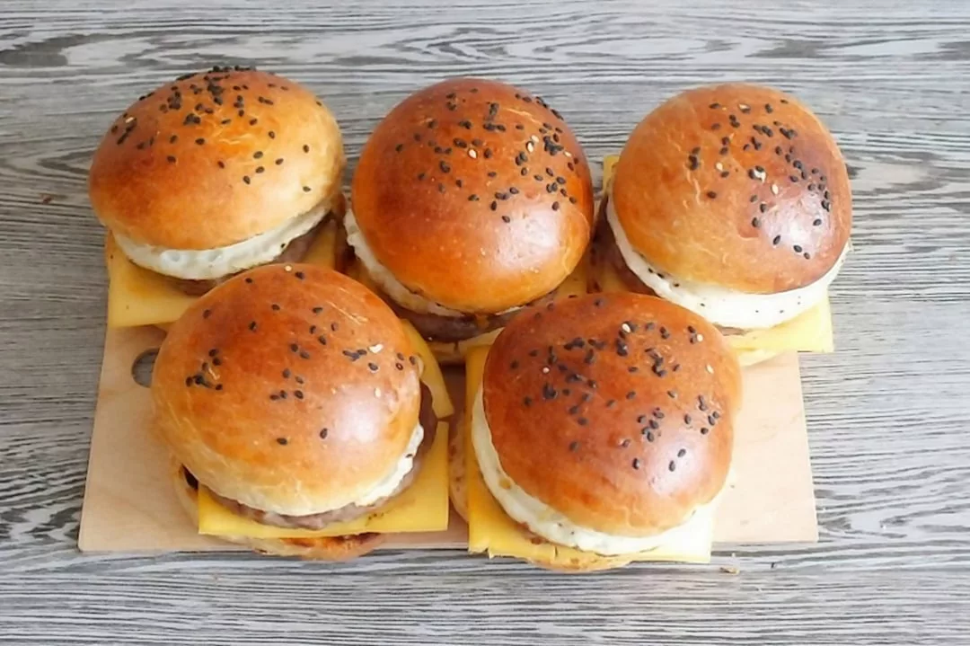 Fünf Cheeseburger mit schwarzen Sesambrötchen und sichtbaren Käsescheiben auf einem Holzbrett angerichtet - perfekt für Spaß in der Küche oder zum Kochen mit Kindern auf einer grauen Holzoberfläche angerichtet.