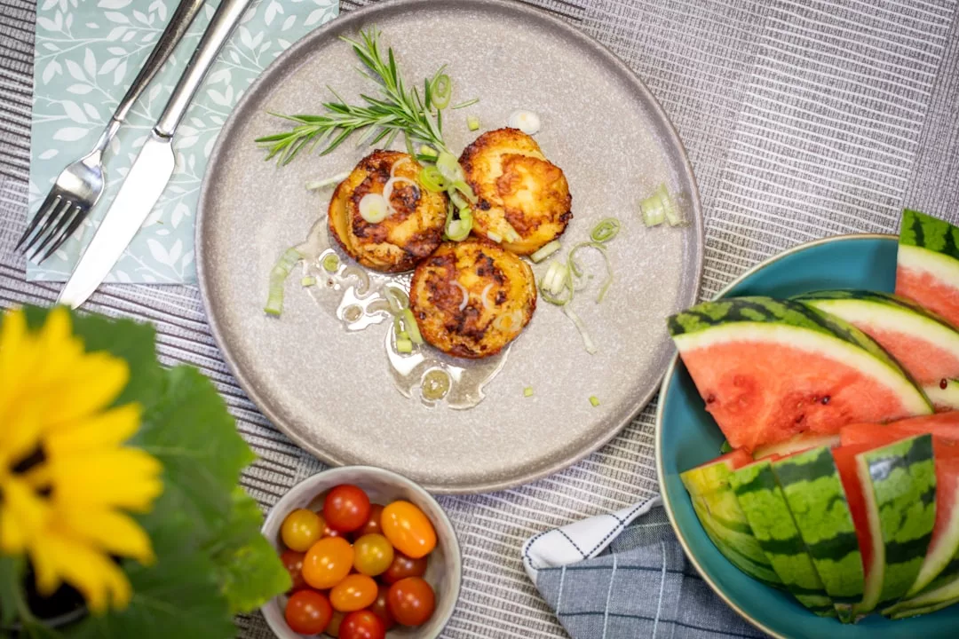 Ein Teller mit drei gegrillten Käsescheiben, garniert mit Frühlingszwiebeln und Rosmarin, zeigt neue Ideen in der Küche, serviert mit einer Schale Kirschtomaten und einem Teller mit Wassermelonenscheiben.