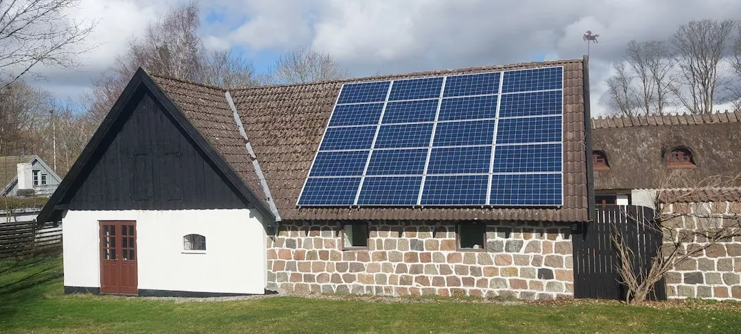 Ein Gebäude mit einem Ziegeldach ist auf einer Seite mit Solaranlagen ausgestattet. Das Gebäude mit seinen steinernen und weiß gestrichenen Wänden ist von Gras und Bäumen umgeben und spiegelt das Engagement für erneuerbare Energie wider.