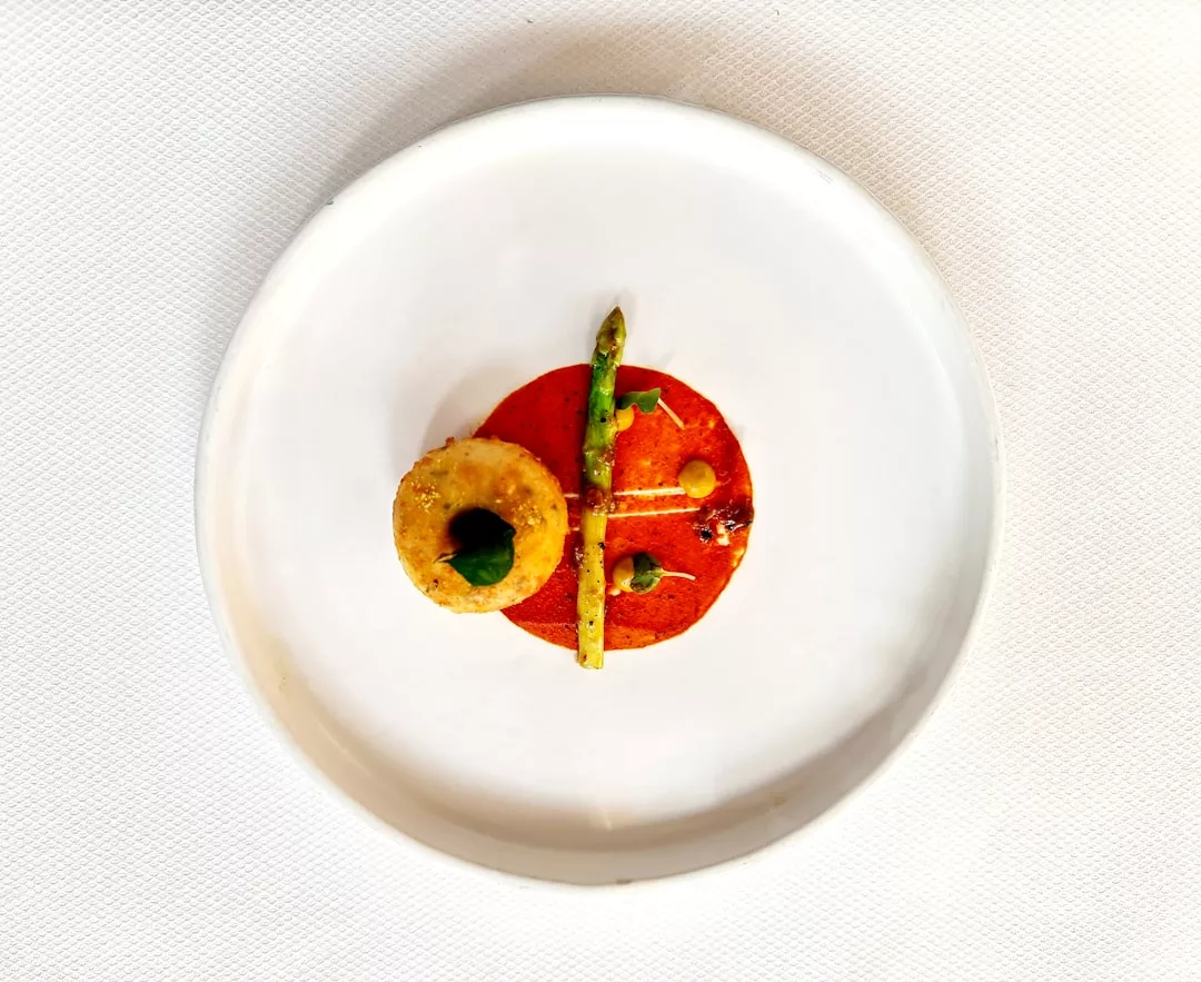 Ein weißer Teller mit einer kreisrunden Portion roter Sauce, einem gebratenen Ring, einer einzelnen Spargelstange und kleinen Garnituren, die fein säuberlich darauf angeordnet sind - eine Inspiration für leckere Alternativen und vegane Gerichte.
