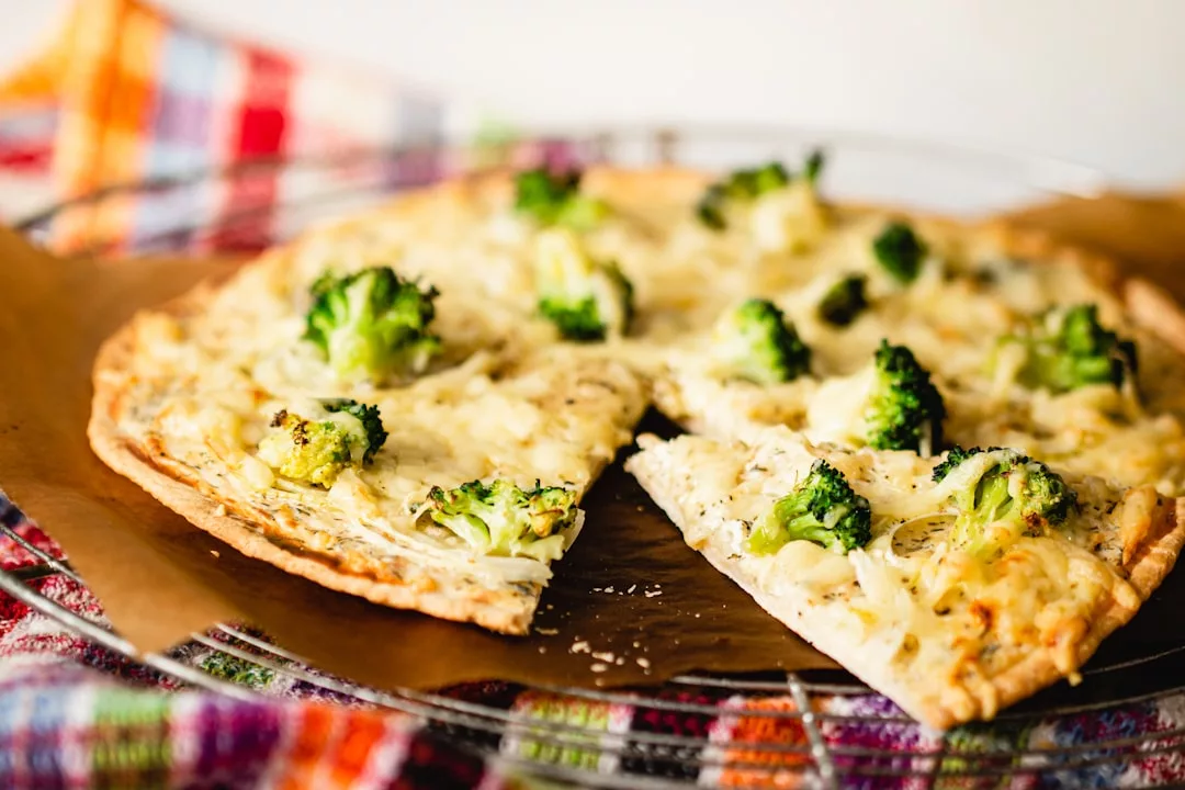 Eine köstliche, mit Brokkoli und Käse belegte weiße Pizza mit dünner Kruste, bei der eine Scheibe leicht vom Rest getrennt ist, liegt auf einem Drahtgestell über einem bunten Tuch - perfekt für Fans vegetarischer Gerichte.