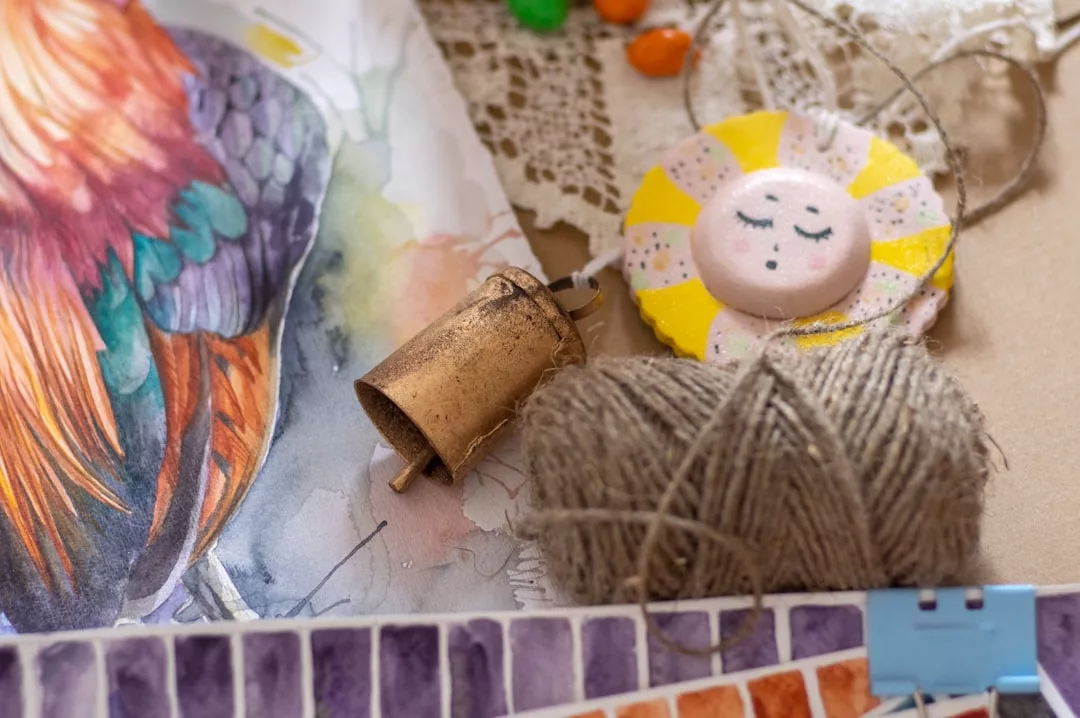 Eine Garnspule, eine kleine Messingglocke, ein gemaltes Sonnenornament, Spitzen und Aquarellfarben liegen auf dem Tisch - perfekte Bastelideen für Kinder, die Basteln und Selbermachen lieben und lebendige Federkunstwerke lieben.