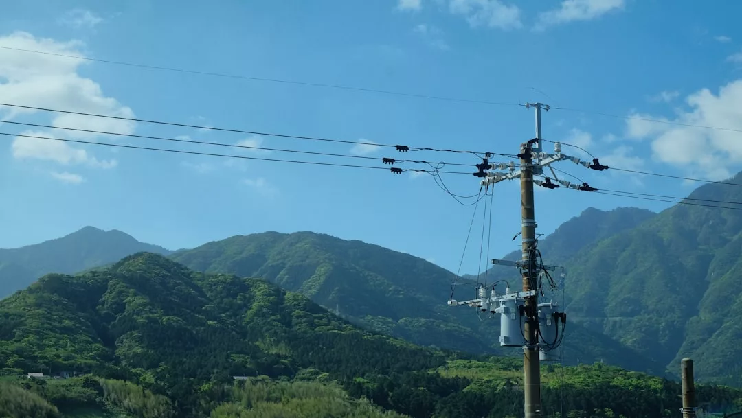 Ein Strommast mit Stromleitungen steht vor grünen, baumbewachsenen Bergen unter einem blauen Himmel mit vereinzelten Wolken, der die Vielfalt und den Genuss naturverbundener Landschaften widerspiegelt und zu Gedanken an kulturelle Veranstaltungen in der Nähe anregt.