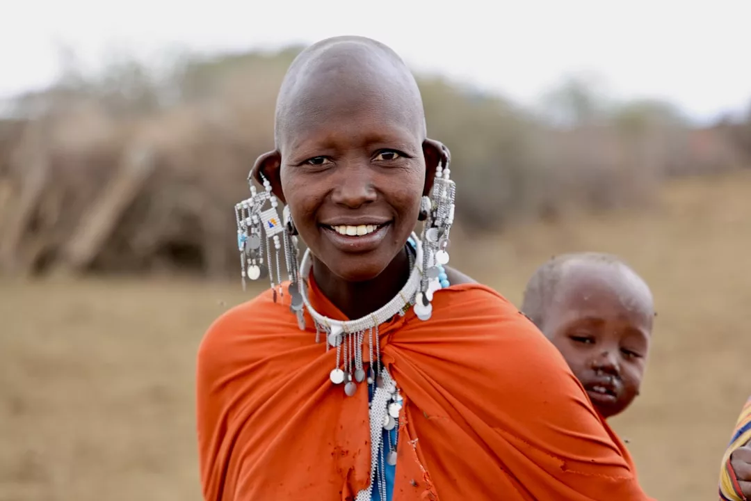 Eine Person, die traditionellen Maasai-Schmuck und orangefarbene Kleidung trägt, lächelt mit einem Kind auf dem Rücken in die Kamera und fängt damit die Schönheit der Welt und das besondere Gefühl ein, bei Kulturreisen neue Menschen zu entdecken.
