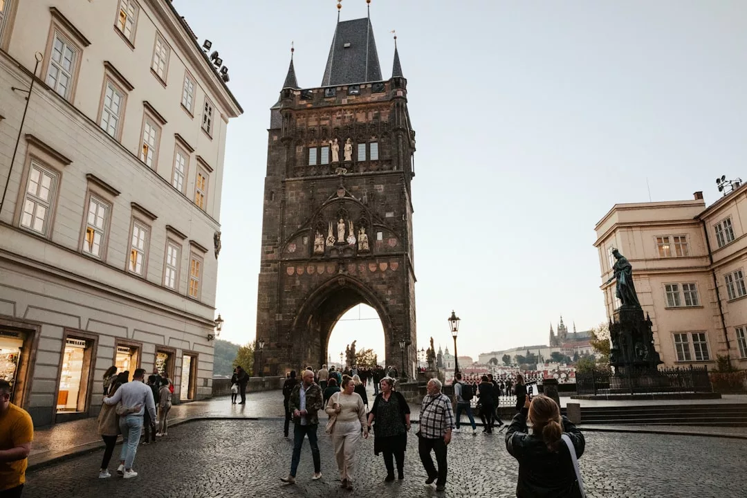 Menschen spazieren in der Nähe eines historischen Steinturms mit einem gewölbten Eingangstor, flankiert von klassischen europäischen Gebäuden, und genießen die reiche europäische Kultur in der Abenddämmerung in dieser städtischen Umgebung.