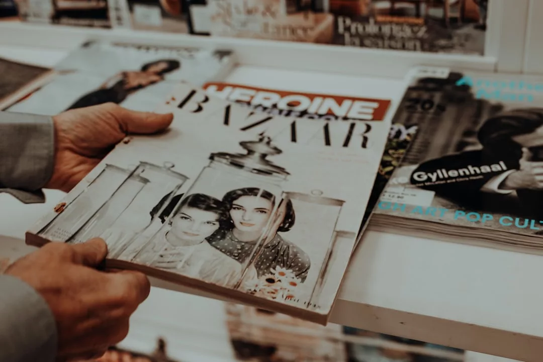 Eine Person hält ein "Harper's Bazaar"-Magazin mit einem schwarz-weißen Titelbild, auf dem zwei Frauen abgebildet sind, neben anderen Kulturzeitschriften in einem Regal, das Vielfalt und kulturelle Reflexion in modernen Printmedien beleuchtet.