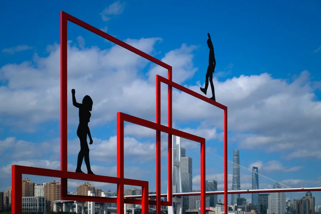 Rote Metallrahmen mit schwarzen Silhouettenfiguren erinnern an Kunst im öffentlichen Raum, vor einer Stadtlandschaft mit Hochhäusern und einem teilweise bewölkten Himmel.