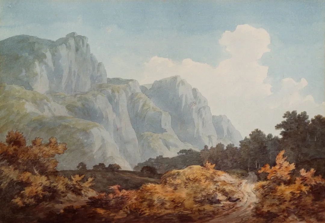 Ein gewundener Feldweg führt durch herbstliches Laub zu hohen, schroffen Bergen unter einem teilweise bewölkten Himmel und lädt zu einer formalen Betrachtung von Natur und Landschaft in der modernen Welt ein.