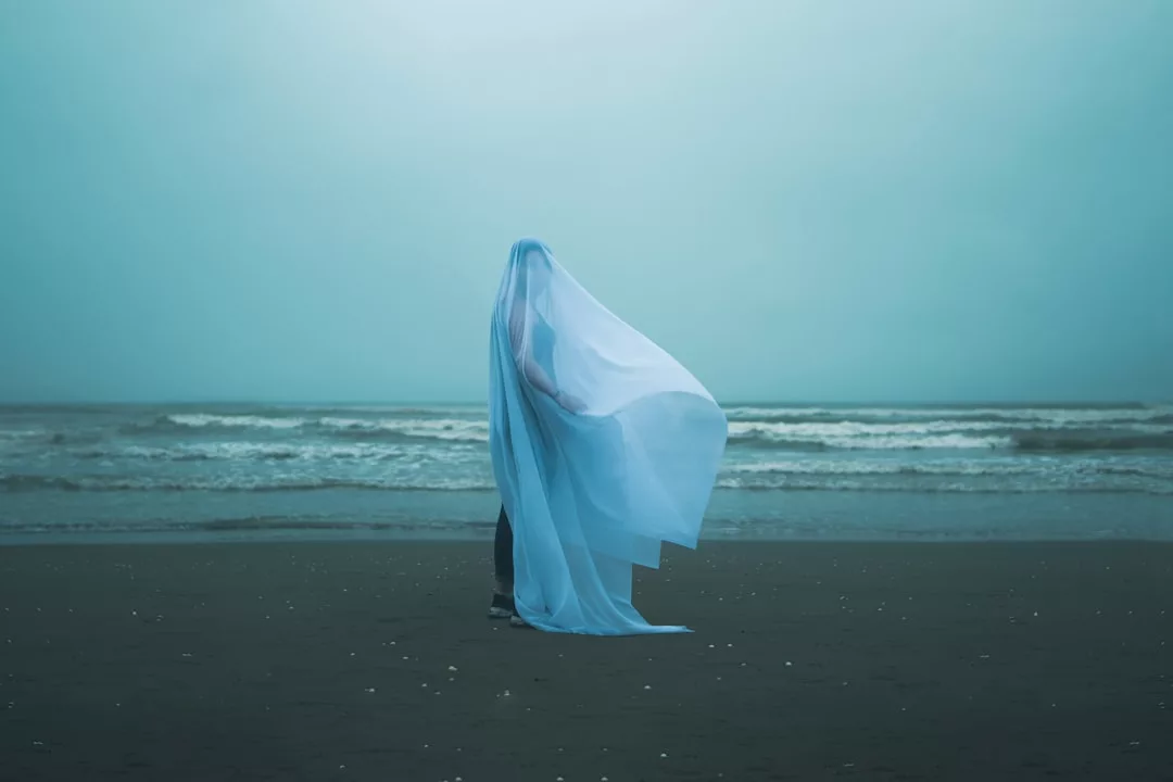 Eine Person steht am Strand, eingehüllt in ein fließendes hellblaues Laken, und blickt unter einem bedeckten Himmel auf das Meer - ein heiterer Moment der Bildkunst, der pure Ästhetik einfängt.