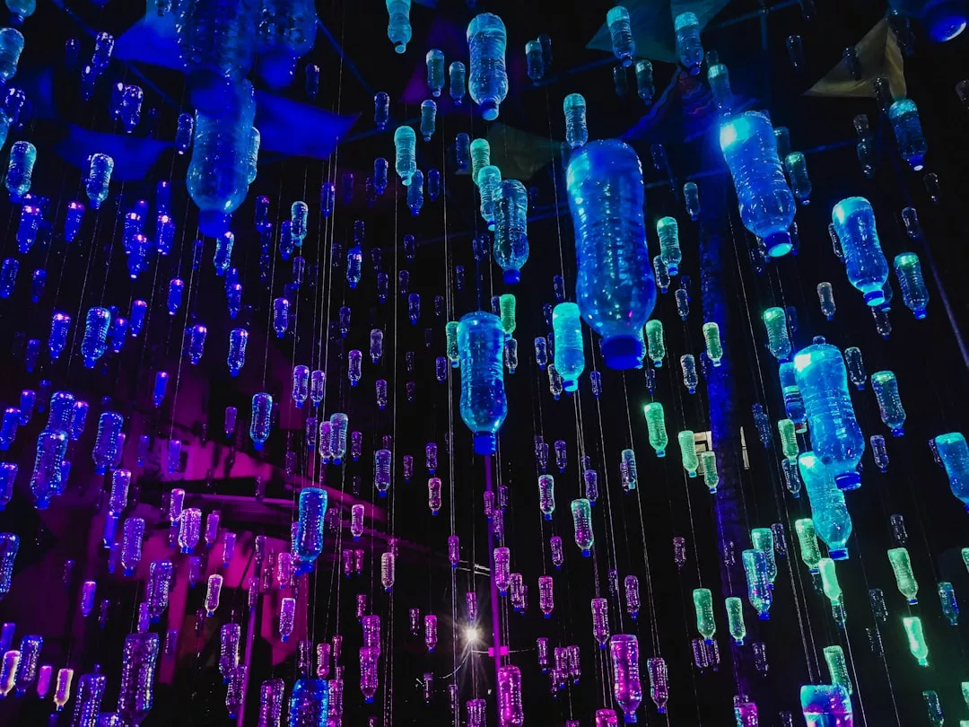 Mehrere Plastikflaschen hängen von der Decke und werden von blauen, violetten und grünen Lichtern in einer dunklen Umgebung beleuchtet - ein Beispiel für künstlerische Innovation, die formale Ästhetik in zeitgenössischen Kunstinstallationen erforscht.