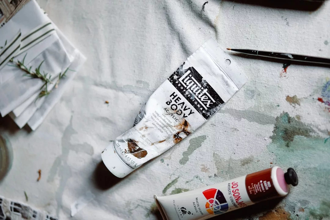 Eine Tube Liquitex Heavy Body Acrylic Paint, eine rote Farbtube und ein Pinsel liegen auf einer fleckigen, mit Farbe bespritzten Oberfläche - perfekte Werkzeuge für die kreative Schöpfung in Künstlerateliers.
