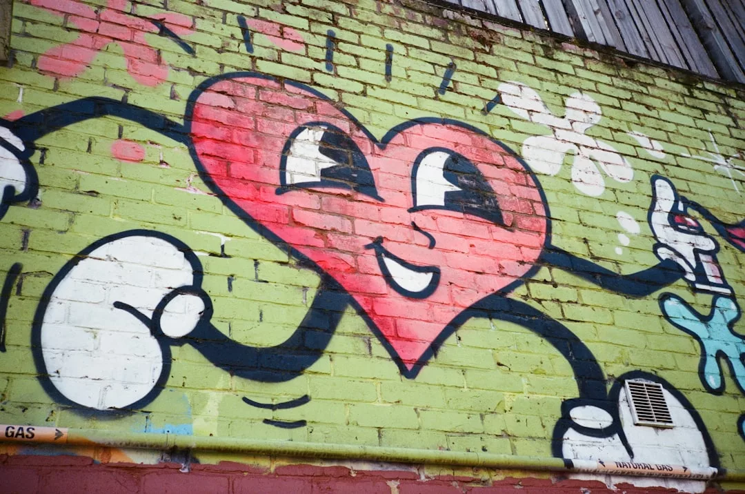 Das Graffiti eines lächelnden Cartoon-Herzens mit Armen und Beinen auf einer grünen Backsteinmauer, umgeben von gemalten Blumen und Formen, zelebriert die Vielfalt der Kunst und kreative künstlerische Ausdrucksformen.