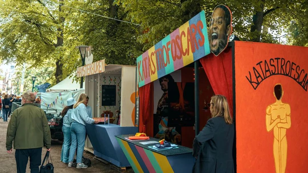 Bei einer Veranstaltung unter freiem Himmel laufen die Menschen an farbenfrohen Karnevalsständen mit verspielten Schildern unter Bäumen vorbei, die die lebhaften Trends der Kunstwelt widerspiegeln. Ein Stand zeigt einen großen Ausschnitt eines Gesichts und die Aufschrift "KATASTROOSCARS".