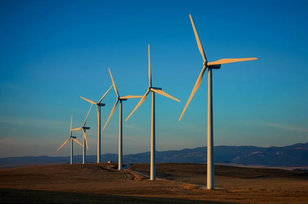 Fünf große Windturbinen stehen in einer Reihe auf einer grasbewachsenen Ebene mit Bergen im Hintergrund und präsentieren erneuerbare Energien unter einem klaren blauen Himmel.