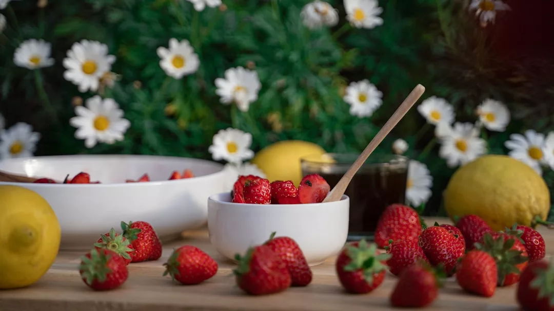 Eine weiße Schale mit Erdbeeren und einem Holzlöffel steht auf einem Tisch neben Zitronen, weiteren Erdbeeren und einer weiteren Schale, mit Gänseblümchen und Grünzeug im Hintergrund - perfekt für leckere Rezepte und frische Mittagessen Rezepte.