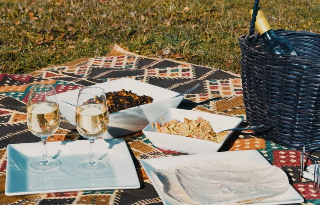 Ein Picknick-Set mit zwei Gläsern Weißwein, Tellern mit Essen, Fladenbrot und einem Korb mit einer Weinflasche auf einer gemusterten Decke im Freien - perfekt für einen perfekten Tag mit köstlichen Picknick-Rezepten.