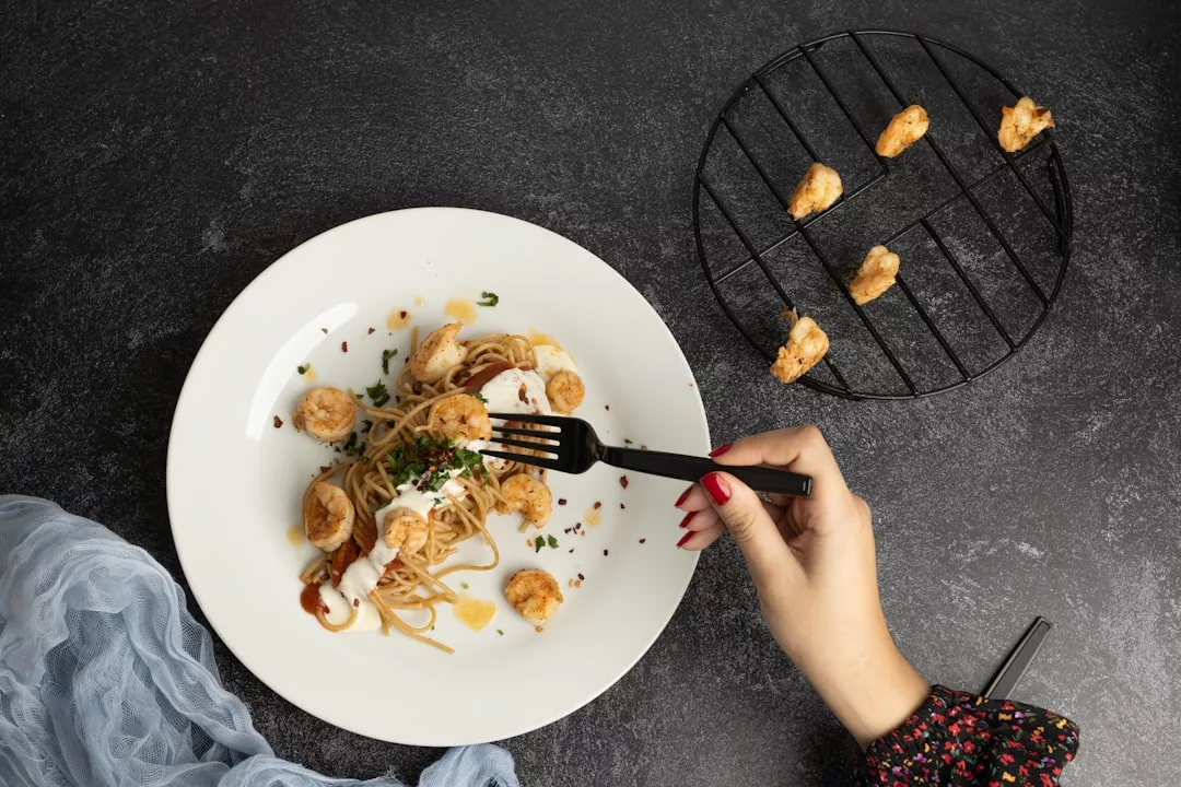 Eine Hand mit einer Gabel ist im Begriff, Krabbenpasta auf einem weißen Teller zu essen; mehrere Krabben liegen auch auf einem schwarzen Kühlregal in der Nähe - perfekt für leckere Rezepte und Rezepte für jeden Tag.