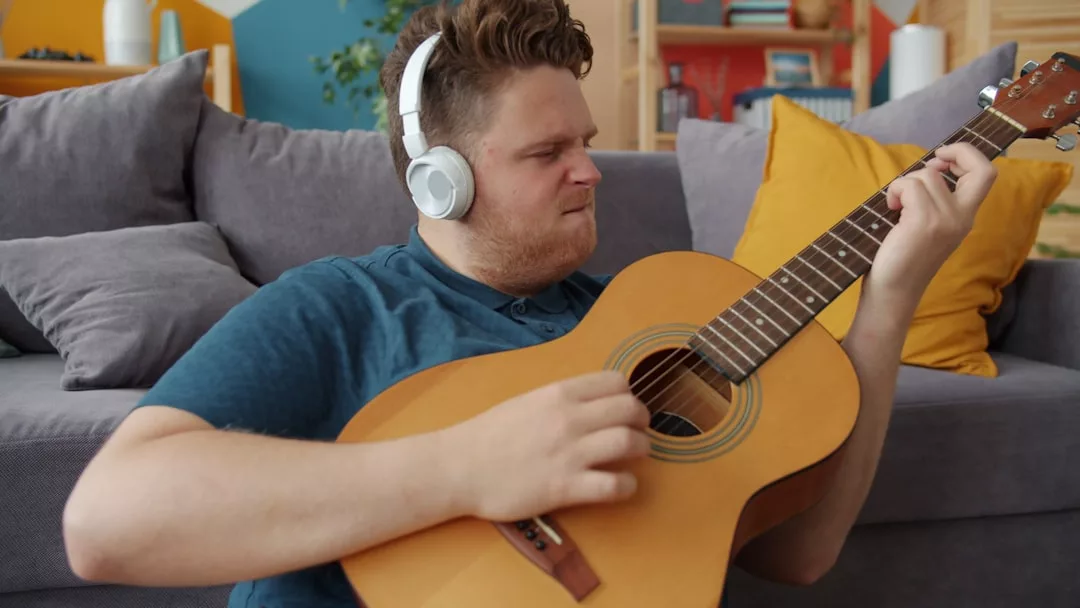 Eine Person mit Kopfhörern sitzt auf dem Boden und konzentriert sich auf das Spielen einer akustischen Gitarre. Im Hintergrund steht ein graues Sofa mit gelben Kissen - perfekt für alle, die nach Tipps Musikinstrument oder nach Anleitung Musikinstrument Inspiration suchen.
