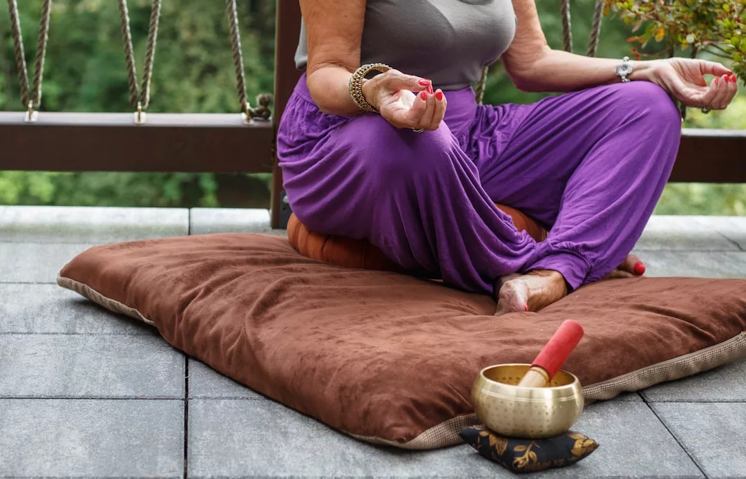 Eine Person in lila Hosen sitzt im Schneidersitz auf einem Kissen und meditiert im Freien mit einer Klangschale und einem Schlegel in der Nähe - eine ideale Szene für Anfänger, die Meditationstechniken erkunden wollen.