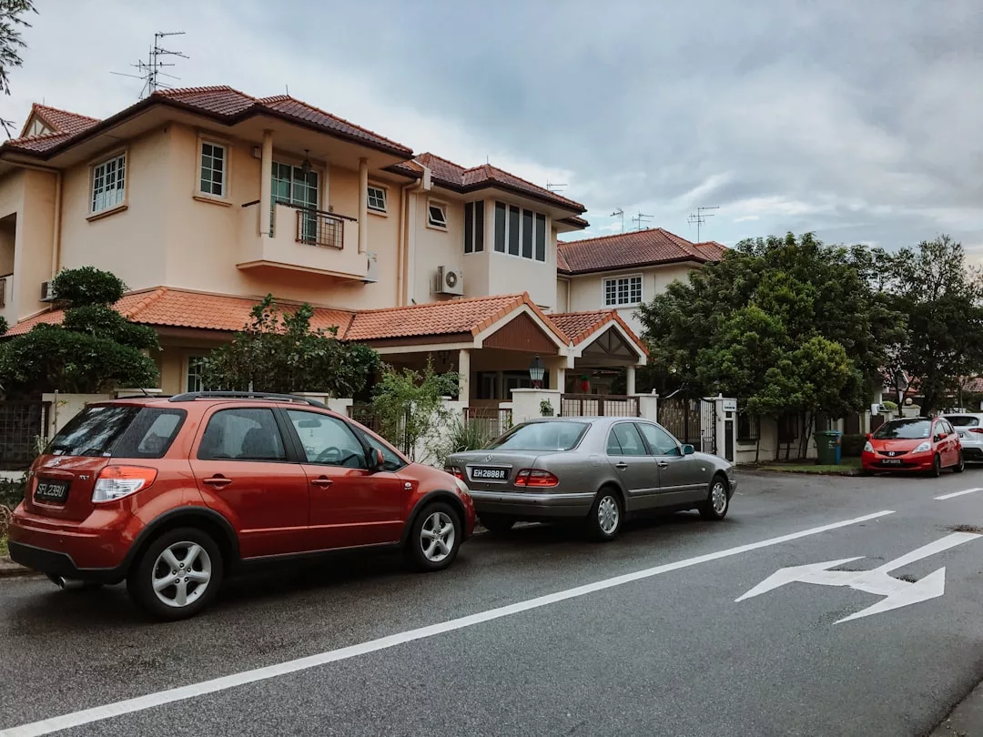 Drei Autos parken in einer Wohnstraße neben einem beigen, zweistöckigen Haus mit rotem Ziegeldach und umlaufender Begrünung unter einem wolkenverhangenen Himmel - ideal, um Mietwagen-Tipps auszutauschen oder Mietwagen-Empfehlungen zu diskutieren.