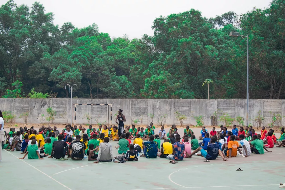 Eine große Gruppe von Menschen, vor allem Kinder in bunten Hemden, sitzt auf einem Sportplatz im Freien und blickt auf einen Redner, der über Sportmotivation spricht, während im Hintergrund Bäume und eine Betonmauer zu sehen sind.