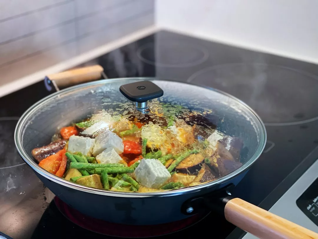 Eine Pfanne mit Deckel gart gemischtes Gemüse und Tofu auf einem Elektroherd, wobei der Dampf am Glasdeckel sichtbar ist - ein Beispiel für umweltfreundliches Kochen und nachhaltiges Kochen.