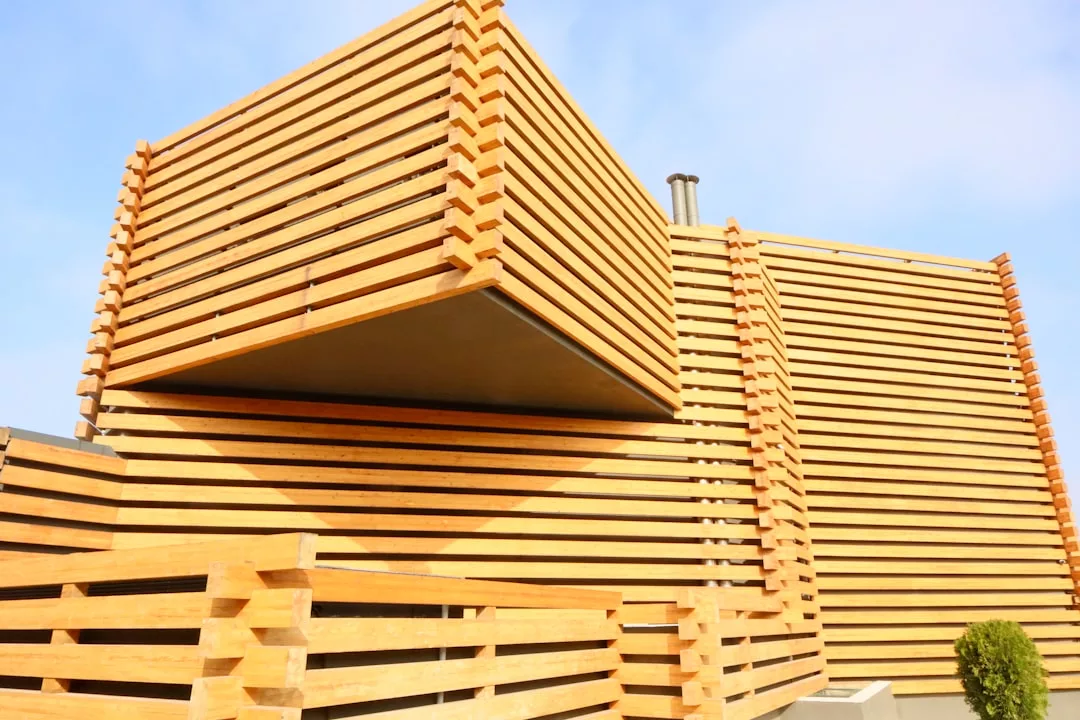 Modernes Gebäude mit kantigen, sich überschneidenden Abschnitten aus nachhaltigen Baustoffen mit hellen horizontalen Holzlatten und einem Zaun vor blauem Himmel.