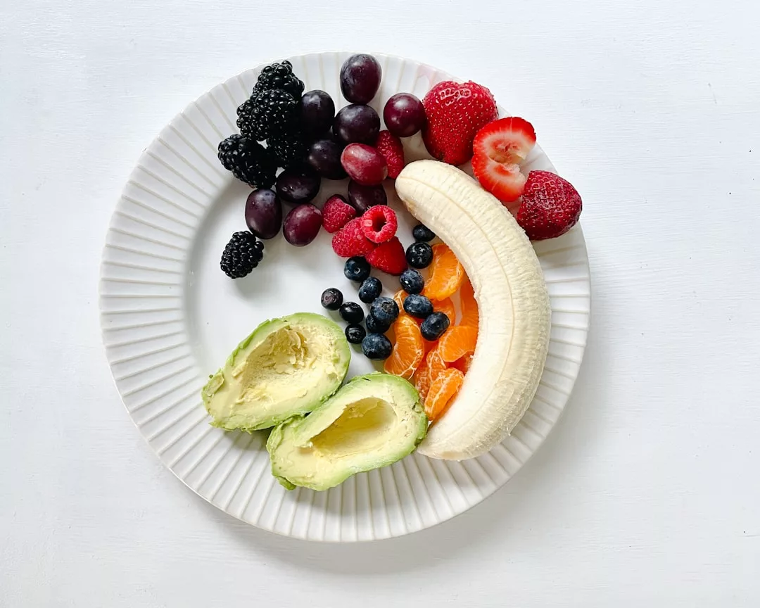 Ein weißer Teller mit Avocadohälften, einer geschälten Banane, Orangenscheiben, Erdbeeren, Brombeeren, Himbeeren, Heidelbeeren und Weintrauben auf weißem Hintergrund - eine Auswahl nährstoffreicher Lebensmittel voller Vitamine und Mineralstoffe.