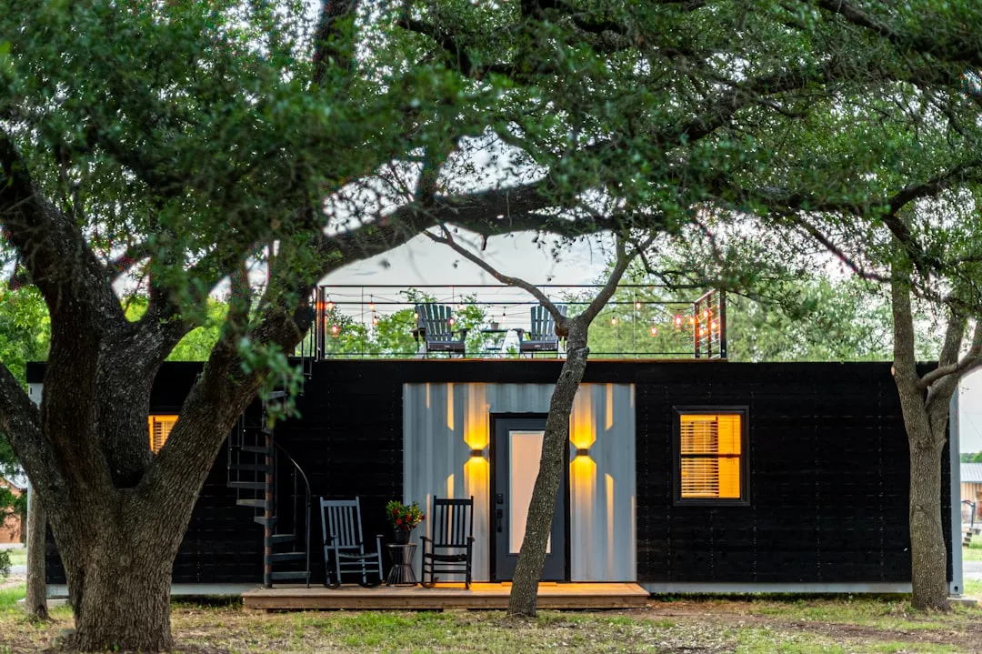 Das moderne kleine Haus aus einem Schiffscontainer verfügt über eine Dachterrasse, eine kleine Veranda mit zwei Schaukelstühlen und ist von großen Bäumen umgeben - perfekt für alle, die Tipps zum autarken Leben oder Off-Grid-Leben in der Natur suchen.