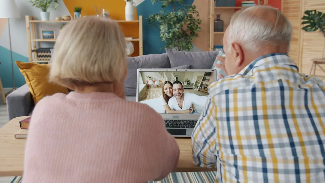 Zwei ältere Menschen sitzen auf einer Couch und genießen die Familienkommunikation, während sie in einem gemütlichen, farbenfrohen Wohnzimmer einen Videogespräch mit einem lächelnden Paar führen, das auf einem Laptop-Bildschirm angezeigt wird.