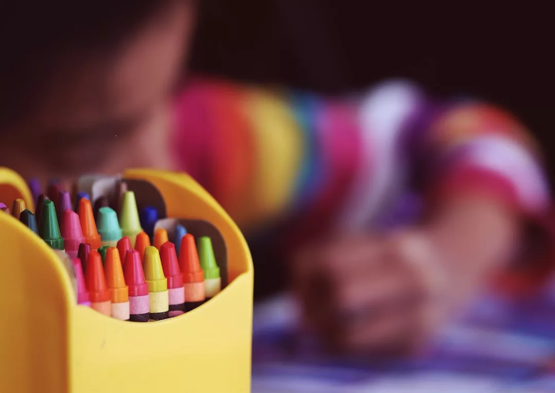 Eine gelbe Schachtel mit bunten Buntstiften steht im Mittelpunkt, während ein Kind in einem regenbogengestreiften Hemd den unscharfen Hintergrund bunt färbt - ein perfekter Schnappschuss kreativer Bildung zu Hause.