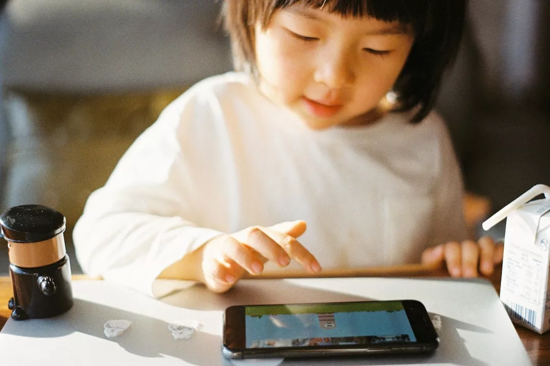 Ein junges Kind benutzt ein Smartphone auf einem Tisch, um pädagogische Apps zu erforschen. In der Nähe stehen ein kleiner schwarzer Behälter, zwei Steine und ein Karton mit Getränken.