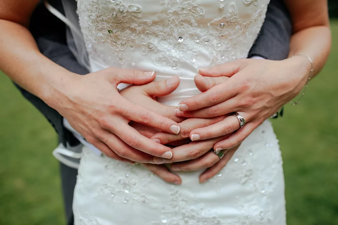 Nahaufnahme einer Person in einem weißen Hochzeitskleid, die von hinten umarmt wird, was Partnerschaften symbolisiert, wobei sich die Hände beider Personen auf dem Kleid überlappen.