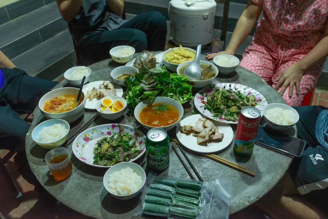 Ein runder Tisch, gedeckt mit Schüsseln mit Reis, Tellern mit Fleisch, Gemüse, Suppe, Dosengetränken und drei Personen, die darum herum sitzen - die perfekte Partyfood-Inspiration mit köstlichen Rezepten und Tipps für Ihre nächste Zusammenkunft.