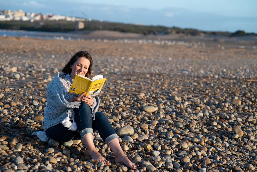 Eine Frau sitzt barfuß an einem felsigen Strand und liest ein gelbes Buch über Selbstentwicklung, während im Hintergrund Gebäude und Bäume zu sehen sind.