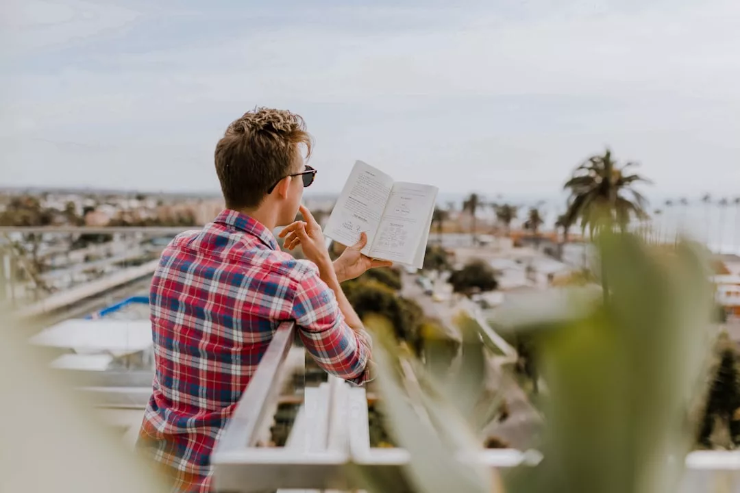 Eine Person in einem karierten Hemd liest ein Buch über Persönlichkeitsentwicklung, während sie sich an ein Balkongeländer lehnt, mit einer Stadtlandschaft und Palmen im Hintergrund - ein ruhiger Moment der Persönlichkeitsentwicklung.