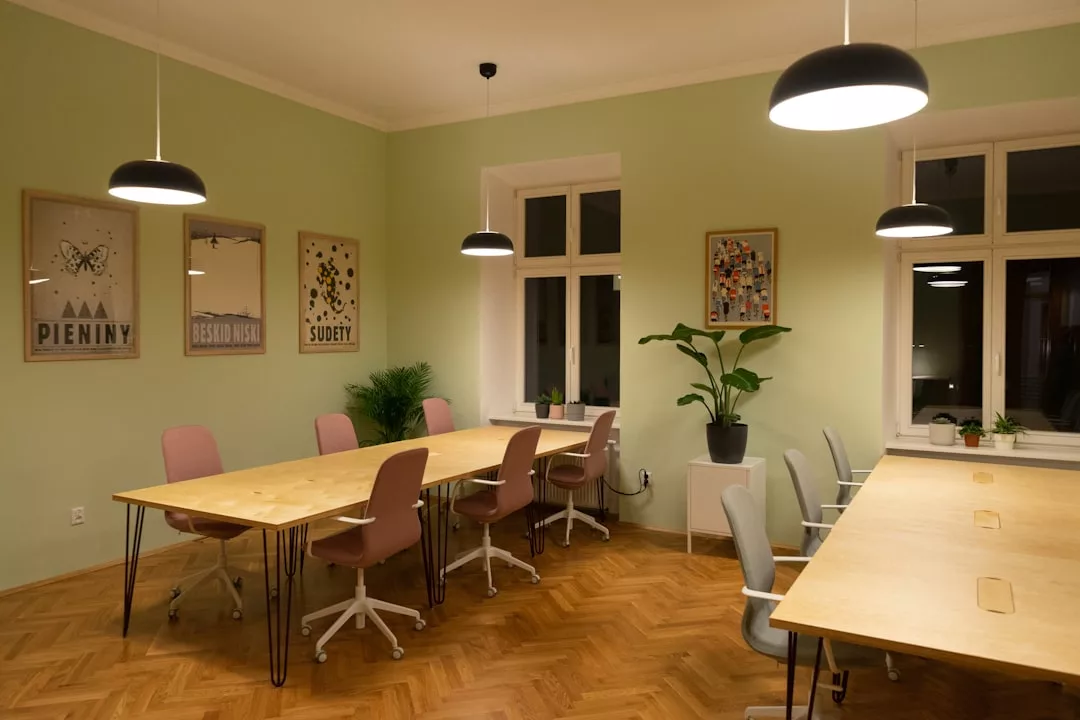Ein moderner Büroraum mit Holzschreibtischen, rosa und grauen Stühlen, Topfpflanzen und gerahmten Kunstwerken an pastellgrünen Wänden, beleuchtet von Deckenleuchten - ein Ergebnis sorgfältiger Planung und energieeffizienter Renovierung.