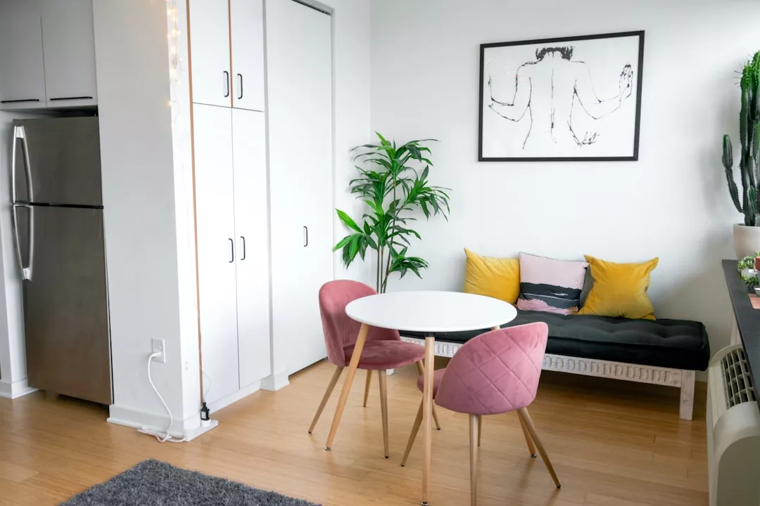 Eine kleine Essecke mit einem runden weißen Tisch, zwei rosafarbenen Stühlen und einer schwarz-weißen Couch mit gelben Kissen zeigt die clevere Raumgestaltung für kleine Wohnungen, komplett mit einer Topfpflanze und einem Kühlschrank in dieser modernen Wohnung.