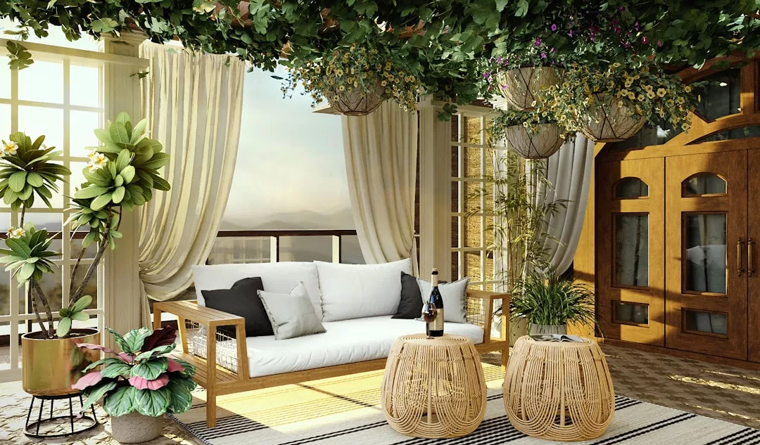 Eine sonnenbeschienene Terrasse mit einem Holzsofa, weißen Kissen, zwei Korbhockern, Topfpflanzen, hängenden Grünpflanzen und großen Fenstern mit cremefarbenen Vorhängen - die perfekten Raumgestaltungsideen für ein stilvolles Zuhause.