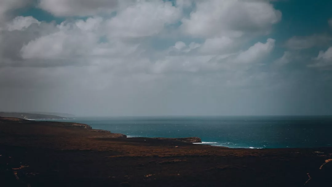 Ein wolkenverhangener Himmel über einer dunklen Küstenlinie, an der sich die Wellen brechen und das Meer sich bis zum Horizont erstreckt - die perfekte Inspiration für Reisetipps, wenn Sie stressfrei entlang malerischer Küstenrouten reisen möchten.
