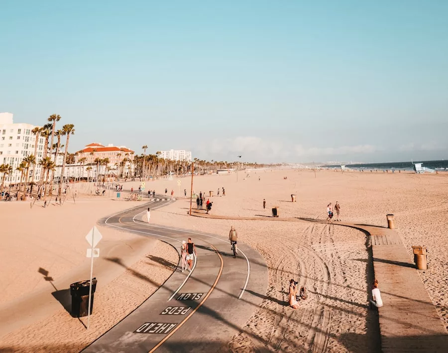 Ein breiter Strand mit einem gepflasterten Radweg, ein paar Spaziergänger und Radfahrer, Palmen und Gebäude im Hintergrund unter einem klaren blauen Himmel - das erinnert Sie daran, dass eine Reiseversicherung für solche unbeschwerten Reisemomente niemals überflüssig ist.