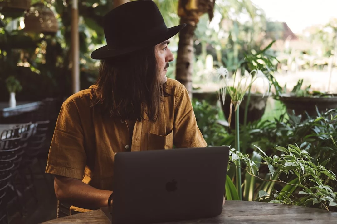 Eine Person mit braunem Hemd und schwarzem Hut sitzt an einem Tisch im Freien mit einem Laptop und blickt zur Seite, umgeben von üppigen grünen Pflanzen - sie ist auf der Suche nach einer besseren Work-Life-Balance und setzt auf Fernarbeit.