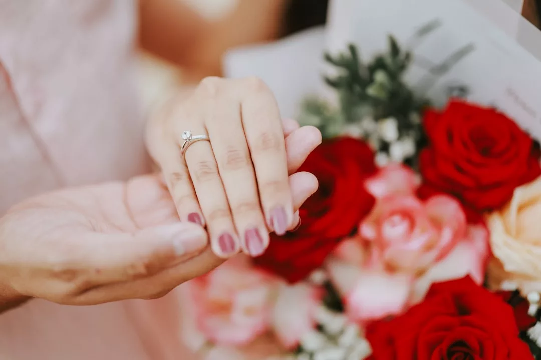 Nahaufnahme einer Hand, die einen Verlobungsring trägt, mit einer anderen Hand, die ihn hält, und einem Strauß roter und rosa Rosen im Hintergrund - ein Moment voller Liebe und romantischer Überraschungen.