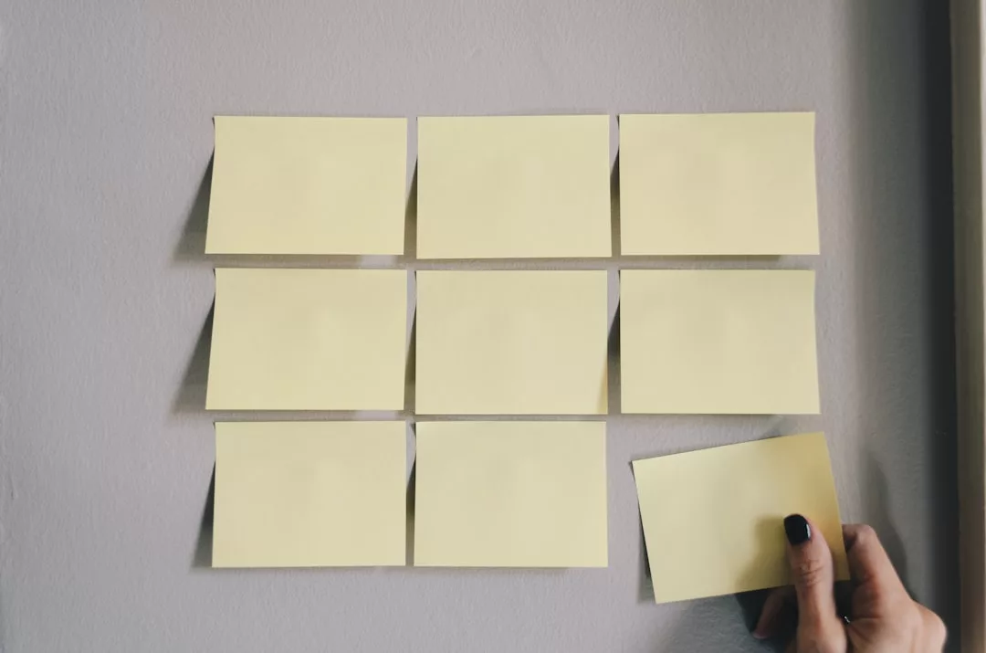 Neun gelbe Haftnotizen sind in einem 3x3-Raster auf einer grauen Wand angeordnet, wobei eine Hand die untere rechte Notiz platziert oder entfernt - ein visueller Hinweis für effektive Strukturierung und Selbstorganisation von Arbeitsprozessen.