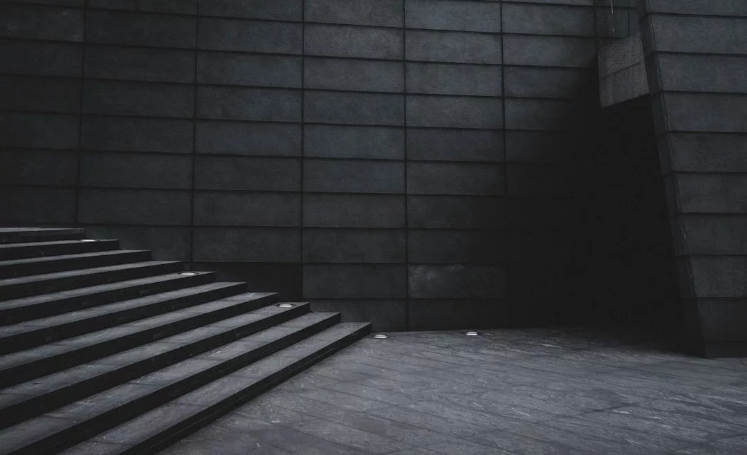 Eine breite Betontreppe führt links neben einer glatten, dunkel gefliesten Wand hinauf in eine schattige Ecke; die Szene ist leer und von weichem, gedämpftem Licht erhellt - subtile Kunst in Selbstdarstellung und Form.