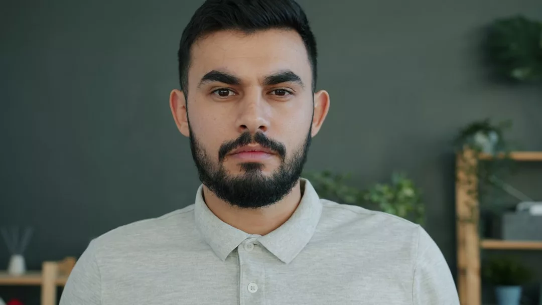 Ein Mann mit dunklem Haar und Bart, der ein hellgraues Poloshirt trägt, steht in einem Haus und blickt mit neutralem Blick in die Kamera. Im Hintergrund sind Regale und Pflanzen zu sehen - perfekt für Tipps, wie du Selbstvertrauen aufbauen und Selbstsicherheit stärken kannst.
