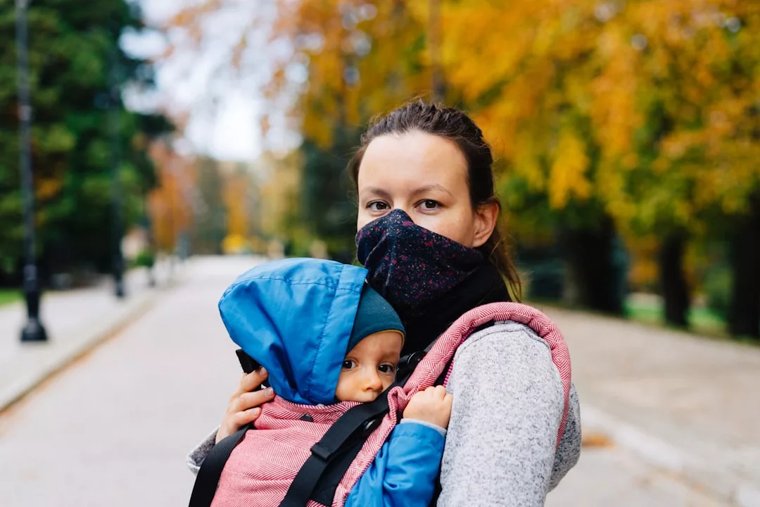 Eine Frau mit Gesichtsmaske trägt ein Baby in einer Babytrage, während sie auf einem Weg in einem Park mit Herbstlaub steht, um zu zeigen, wie wichtig die Kindersicherheit auch im Freien ist.